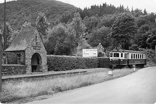 Das Bild zeigt einen ruhigen Bahnhof in einer l ndlichen Umgebung, aufgenommen in Schwarz‑Wei . Im Vordergrund verl uft ein schmaler Stra en- oder Wegabschnitt, parallel dazu links ein einzelnes Bahngleis. Links im Bild steht ein kleines, einschiffiges Warteh uschen aus grobem Bruchstein mit einem spitz zulaufenden, geschindelten Dach und einer halbkreisf rmigen Durchgangs ffnung; an der Seitenwand sind grobe Steinfugen gut sichtbar. Hinter dem H uschen befindet sich eine dichte Hecke, dar ber B ume und im Hintergrund ein bewaldeter H gel.   Rechts auf der Fahrbahn bzw. am Gleisrand steht ein einzelner Triebwagen mit geradem Wagenkasten, zwei Fensterreihen und einem Frontbereich mit Scheinwerfern. Der Triebwagen ist etwas zur ckgesetzt vom Betrachter und nach links ausgerichtet. Auf der Seite des Wagens ist eine Beschriftung zu erkennen (Lesbarkeit eingeschr nkt durch die Bildqualit t). Vor und hinter dem Gleis wachsen hohe Grasb schel und Wiesenrandvegetation.    Die Bildstimmung wirkt ruhig und zeittypisch, das Schwarz‑Wei  verleiht der Szene einen historischen Charakter. Es sind keine Personen in Nahaufnahme zu sehen; einzelne Details wie das Dach des Warteh uschens, die Struktur der Hecke und die Form des Triebwagens sind klar erkennbar.