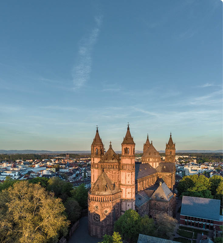 Luftaufnahme des Wormser Doms mit seinen markanten T rmen im warmen Abendlicht. Die gro e romanische Kirche steht zentral und ist von B umen sowie der umliegenden Stadt mit H usern und Stra en umgeben. Im Hintergrund erstreckt sich die weite Landschaft bis zum Horizont unter einem klaren Himmel.