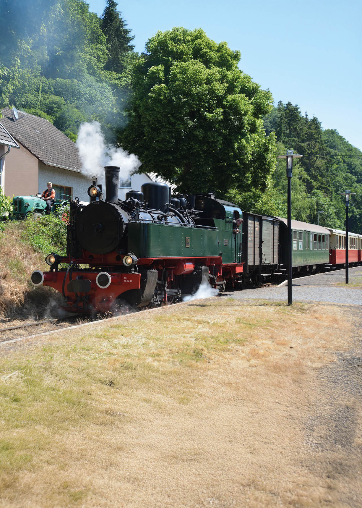 Vorn Mitte im Bild steht eine historische Dampflokomotive auf schmalen Gleisen, leicht nach links geneigt, so dass die Vorderseite zum Betrachter zeigt. Die Lokomotive ist dunkelgr n lackiert, der Rahmen und die vorderen Puffer sind rot, an der Front sind zwei runde wei e Pufferumrandungen sichtbar. Aus dem Schornstein und unter dem Fahrgestell steigt wei er Dampf auf. Hinter der Lok schlie en sich ein geschlossener G terwagen und mehrere Personenwagen in unterschiedlichen Farben (gr n, beige, rot) an, die sich in einer langen Reihe nach rechts hinten fortsetzen.   Links hinter der Lok liegt eine kleine Anh he mit einem hellen, zweigeschossigen Haus und einer kurz geschnittenen B schung; vor dem Haus steht ein geparkter,  lterer gr ner Traktor. Zwei Personen sind seitlich am Hang zu erkennen. Rechts neben den Gleisen verl uft ein gepflasterter Halteplatz mit einer Stra enlaterne; davor erstreckt sich eine Fl che mit trockenem, gelblich-br unlichem Gras. Im Hintergrund wachsen gro e, dichte B ume, insbesondere ein markanter, volumin ser Laubbaum direkt hinter der Lok, der viel Schatten spendet. Weiter hinten schlie en bewaldete H gel die Szene ab.    Die Lichtstimmung ist hell und sonnig, typische Sommeratmosph re. Die Szene wirkt lebendig und nostalgisch, mit deutlichen Kontrasten zwischen der dunklen Lok, dem gr nen Hintergrund und den hellen Wiesenfl chen.    Wichtige Informationen / Kontext:    Motiv: Museumsdampflokomotive mit Zug in kleiner l ndlicher Haltestelle.  Sichtbare Elemente: Dampflok (gr n/rot), Dampfwolken, G ter- und Personenwagen, Gleise, Haus mit Traktor am Hang, gro er Baum, Laterne, trockene Wiese; keine identifizierbaren Gesichtsdaten von Personen.  Stimmung/Zweck: nostalgisch, sommerlich, fahrplan- oder betriebsbezogene Szene (Zug f hrt ab/kommt an).  Empfohlene Bildunterschrift: Museumsdampflok (gr n/rot) mit Wagons verl sst einen kleinen Bahnhof; Dampfwolken steigen auf, Sommerwiese und gro e B ume im Hintergrund.      