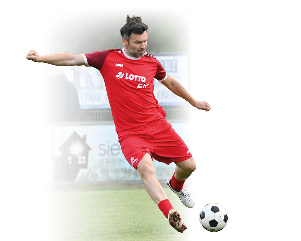 Auf einem gr nen Rasen ist ein Spieler in rotem Trikot in Schu position. Der K rper ist angespannt, der Blick konzentriert auf den schwarz-wei en Ball gerichtet; im Hintergrund ein Zaun mit Werbetafeln und mehrere unscharfe Zuschauer. Die Gesichter der Personen sind nicht erkennbar.