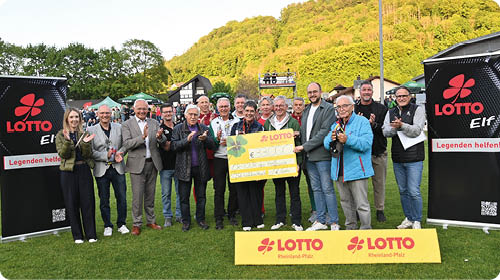 Auf gr nem Rasen posiert eine gemischte Gruppe Erwachsener in einer Reihe; alle blicken zur Kamera und klatschen oder halten applaudierend die H nde. In der Bildmitte tragen zwei Personen gemeinsam einen bergro en gelben Scheck mit der roten Lotto Marke, links und rechts daneben stehen zwei hohe schwarze Rollups mit rotem Kleeblatt Logo und dem Schriftzug „LOTTO“. Im Hintergrund sind weitere Zuschauer, ein H gel mit B umen und mehrere Zelte oder Pavillons zu sehen; die Gesichter der Personen sind nicht erkennbar.