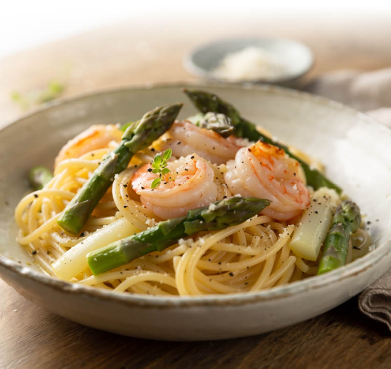 Eine Schale mit Spaghetti steht auf einem Holztisch. Die Pasta ist mit gr nem Spargel und mehreren Garnelen angerichtet und mit geriebenem K se sowie schwarzem Pfeffer bestreut. Im Hintergrund stehen unscharf eine kleine Schale mit K se und ein helles Tuch. Warmes Licht sorgt f r eine appetitliche Stimmung.