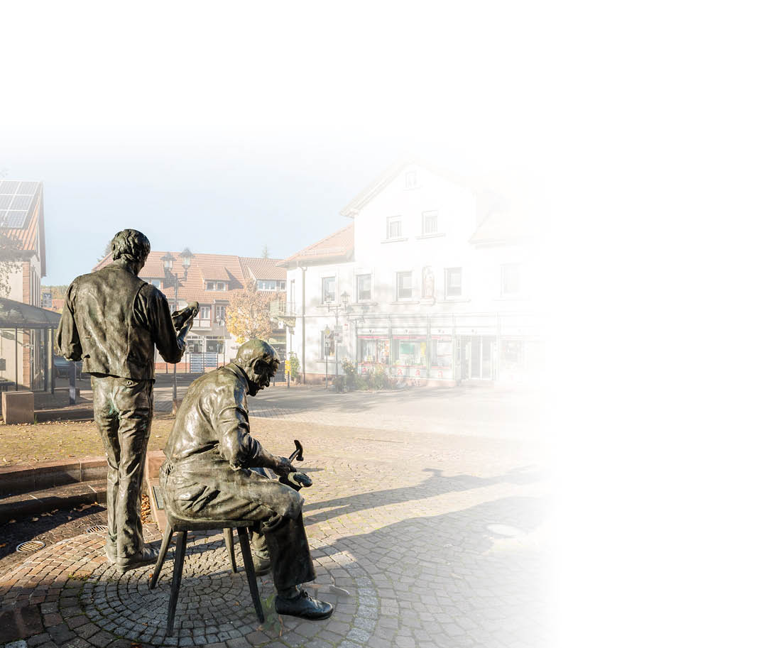Bronzefiguren eines Denkmals auf einem gepflasterten Platz in einer Kleinstadt. Zwei lebensgro e M nner aus Bronze: eine stehende Figur mit dem R cken zur Kamera, die rechte Hand leicht erhoben, und eine sitzende Figur auf einem Stuhl, nach vorn gebeugt mit einem Hammer in der rechten Hand. Beide Figuren tragen Arbeitskleidung; die sitzende Figur hat den Blick auf die H nde gerichtet, als ob sie gerade an einem Objekt arbeitet. Die Figuren stehen auf einem runden Pflasterfeld mit konzentrischen Steinringen; im Hintergrund sind B nke und Geb ude mit Gesch ften und Wohnungen zu erkennen. Die Szene ist sonnig, leichte Schlagschatten liegen auf dem Platz; Stimmung: ruhige, handwerklich gepr gte Stadtszene.