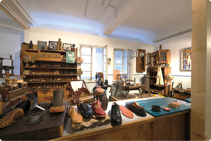Innenansicht einer historischen Schuhmacherwerkstatt, hell ausgeleuchtet durch Fenster an der R ckwand. Im Vordergrund eine lange Arbeitsfl che mit mehreren fertigen Schuhen und Leisten aus Holz: schwarze Lederschuhe, ein roter Damenschuh, ein hellbrauner Halbschuh und mehrere einzelne Leisten. Links stehen weitere Holzleisten und Werkst cke auf einem niedrigen Tisch. Im mittleren und hinteren Bereich ein hoher, dunkler Holzschrank mit vielen kleinen F chern und Schubladen, best ckt mit Lederst cken, Werkzeugen und Musterleisten. Rechts daneben mehrere Arbeitstische mit N hmaschinen, Amboss‑ hnlichen Unterlagen, H mmern und weiteren Schuhmacherwerkzeugen; an der Wand h ngen zus tzliche Werkzeuge und ein Schrank mit Glast r. Die Atmosph re wirkt handwerklich und museal, Materialien dominieren Holz, Leder und Metall.