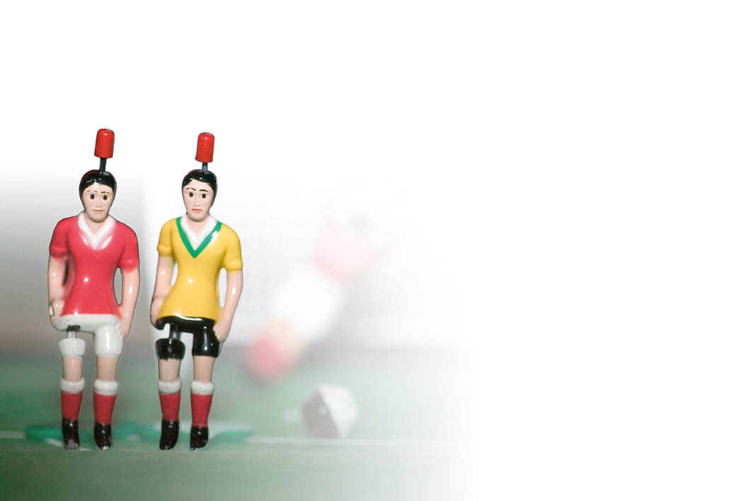 Zwei Tischfu ballfiguren in Fu balltrikot-Pose, frontal, nebeneinander. Links eine Figur mit rotem Trikot, wei en Hosen und roten Kniestr mpfen; rechts eine Figur mit gelbem Trikot, schwarzen Hosen und roten Kniestr mpfen. Beide tragen schwarze Schuhe und haben am Kopf einen roten Zapfen zum Einh ngen in die Stange. Die Gesichter sind nicht detailliert erkennbar. unscharfer, hellgrauer Hintergrund; rechts im Bild ein verschwommener weiterer Spieler. Der Bildausschnitt ist leicht von unten aufgenommen.