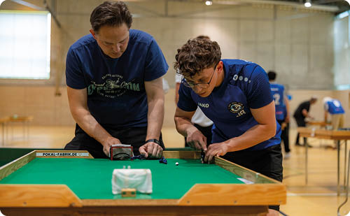 Zwei M nner beugen sich konzentriert ber einen gr nen Spieltisch in einer Halle. Links steht ein Mann mit dunkelblauem T‑Shirt und dunkler Hose, rechts ein j ngerer Mann in blauem Trikot mit Vereinslogo. Beide st tzen sich mit den Oberk rpern nach vorn, die H nde greifen an Spielger te am Tischrand; auf der Tischfl che liegen kleine Spielfiguren und ein wei es Spielobjekt. Im Vordergrund an der Tischkante ist ein kleines Tor oder Beh lter sichtbar. Im unscharfen Hintergrund: weitere Personen und Tische in der Sporthalle, helle W nde und Deckenbeleuchtung. Gesichtsz ge sind nicht erkennbar. Die Stimmung wirkt konzentriert und spielerisch.