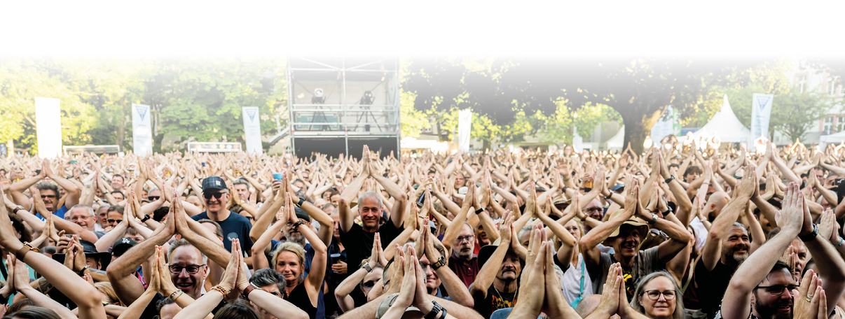 Blick auf ein dichtes Publikum bei einem Open‑Air‑Konzert oder Festival. Hunderte Menschen stehen eng beieinander und f llen das Bild von Vorder- bis Hintergrund. Viele Besucher haben die Arme nach oben gestreckt und klatschen in die H nde; einige halten Smartphones hoch. Die Personen tragen sommerliche, l ssige Kleidung (T‑Shirts, Caps) in unterschiedlichen Farben. Im Hintergrund ist die B hnentechnik und ein Teil der Veranstaltungsfl che zu erkennen, die Szene wirkt lebhaft und energiegeladen. Der Gesamteindruck ist fr hlich und mitrei end, typischer Moment w hrend eines Auftritts, wenn das Publikum gemeinsam applaudiert.