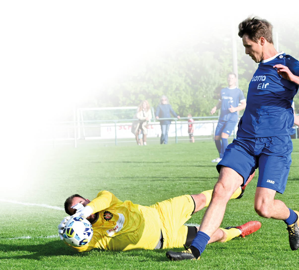 Im Vordergrund rutscht ein Torwart in gelber Spielkleidung mit ausgestreckten Armen ber den gr nen Rasen und f ngt einen Fu ball. Rechts daneben st rzt ein Feldspieler in blauem Trikot und blauen Shorts, er versucht offenbar, den Ball noch zu erreichen.