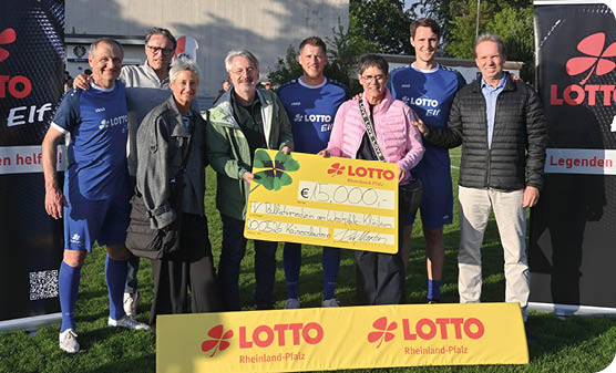 Gruppenfoto auf einer Rasenfl che bei Tageslicht: Neun Personen stehen dicht nebeneinander vor zwei aufgestellten Bannern mit dem Logo „LOTTO“ und einem gelben Werbebanner am Boden. Vier M nner tragen blaue Fu balltrikots und Shorts; drei weitere Erwachsene sind l ssig gekleidet in Alltagsjacken und Hosen, eine Person tr gt eine rosa Jacke. In der Bildmitte halten zwei Personen gemeinsam einen gro en gelben Scheck ber 15.000 Euro mit dem LOTTO-Logo. Im Hintergrund ist ein helles Geb ude und gr nes Buschwerk zu sehen. Die Stimmung wirkt feierlich und freundlich, die Gruppe posiert f r ein offizielles Foto bei einer Scheck bergabe.
