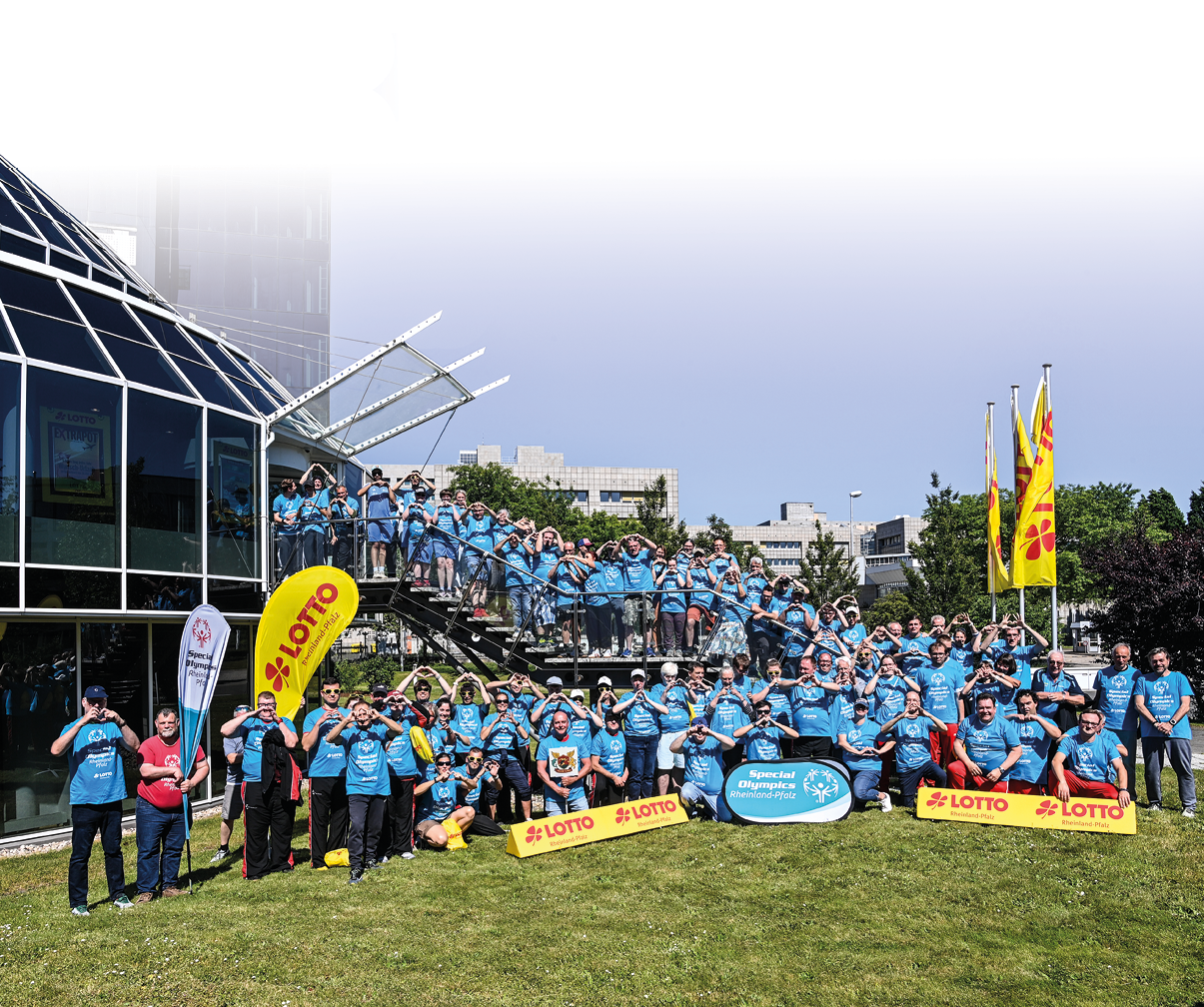 Auf einem sonnigen, wolkenlosen Tag steht eine gro e Gruppe von Menschen in hellblauen T Shirts vor einem modernen Glasgeb ude. Die Gruppe ist ber zwei Ebenen verteilt: ein gro er Teil steht auf einer Au entreppe links, weitere Personen bilden mehrere Reihen auf dem Rasen davor. Vorne liegen zwei gelbe Werbeplattformen mit der Aufschrift „LOTTO“ und ein ovales Schild mit dem Schriftzug „Special Olympics Rheinland Pfalz“. Rechts hinter der Gruppe wehen drei gelbe Fahnen an Masten. Viele der Anwesenden formen mit beiden H nden ein Herzzeichen vor der Brust oder dem Gesicht; einige l cheln in die Kamera. Im Hintergrund sind B robauten und B ume zu sehen. Die Stimmung wirkt fr hlich und gemeinschaftlich.