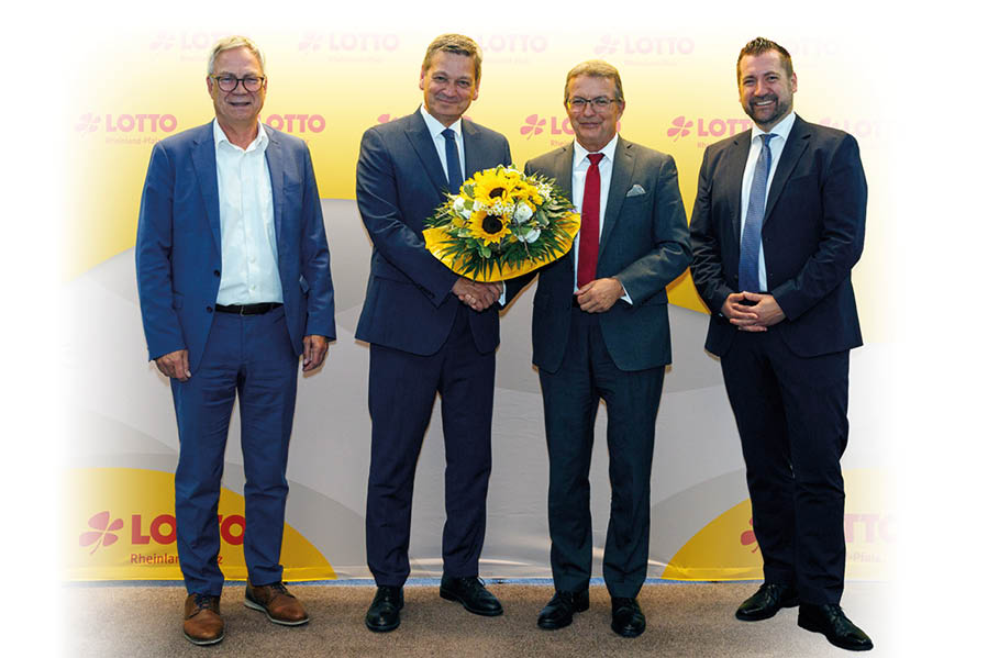 Vier M nner in Anz gen stehen vor einer gelben Wand mit roten „LOTTO Rheinland-Pfalz“-Logos. Die Personen sind dicht nebeneinander aufgestellt; die beiden mittleren halten gemeinsam einen gro en, runden Blumenstrau mit dominierenden Sonnenblumen, gelben und wei en Bl ten sowie gr ner Blattf llung. Alle tragen dunkle Anz ge, Hemden und Krawatten (die zweite Person von rechts mit roter Krawatte), Schuhwerk sind dunkle Business-Schuhe; die  u eren M nner tragen hellere Anz ge bzw. blaue Kleidung. Der Hintergrund ist hellgelb mit dem wiederkehrenden Logo, die Szene wirkt formell und fe.stlich