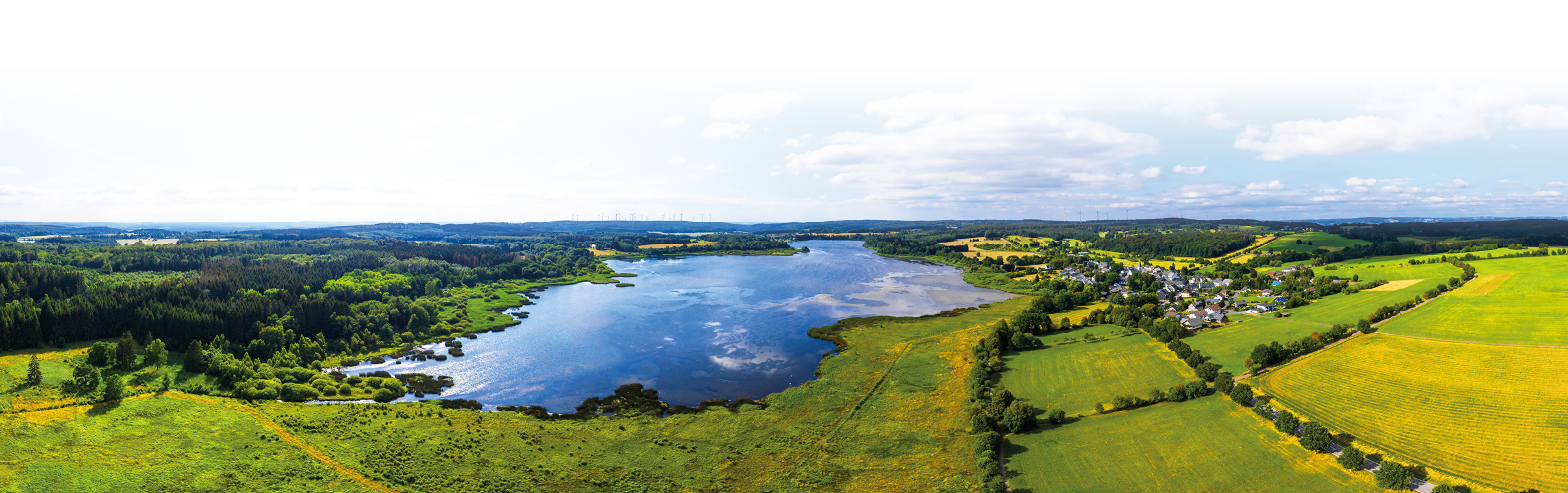 Weitwinkel-Panorama ber eine Seenlandschaft und umliegende Felder an einem hellen, teils wolkigen Tag. Mittelpunkt ist ein langgestreckter, ruhiger See mit spiegelnder Oberfl che, die in der Bildmitte Lichtreflexe zeigt. Links schlie en dichte, dunkelgr ne W lder an das Ufer an, rechts geht das Ufer in offene, hellgr ne und gelblich get nte Wiesen und Ackerfl chen  ber. Am rechten Bildrand liegt ein kleines Dorf mit mehreren Geb uden und Stra en, die sich zwischen Feldern und Baumgruppen schl ngeln. Am fernen Horizont sind weitere H gel und vereinzelt Windr der zu erkennen. Der Himmel nimmt das obere Bilddrittel ein, ist  berwiegend blau mit gr  eren wei en Wolkenfeldern. Stimmung: weitl ufig, ruhig und sonnig mit l ndlichem Charakter.