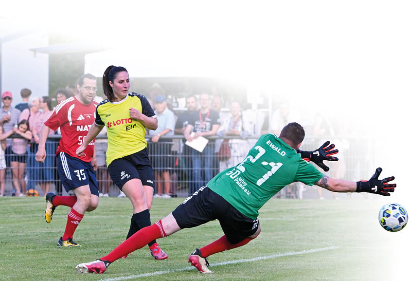 Nahaufnahme einer lebhaften Spielsituation auf einem Fu ballfeld. Im Vordergrund streckt ein Torwart in gr nem Trikot mit der Nummer 31 sich seitlich aus, beide Beine weit gespreizt, und versucht, den Ball zu erreichen. Der Ball liegt rechts im Bild knapp vor seinem ausgestreckten Fu . Vor dem Torwart dr ngen zwei Feldspieler dicht nebeneinander: eine Spielerin in gelbem Trainingshemd mit schwarzer Hose bewegt sich Richtung Ball, links daneben ein Spieler in rotem Trikot mit blauen Hosen. Im Hintergrund hinter der Spielfeldbegrenzung stehen mehrere Zuschauer an einem Zaun, einige unter einem wei en Markt- oder Cateringzelt, die Szene wirkt angespannt und konzentriert. Farbeindr cke: gr ne Spielfl che, helle Trikots in Gelb, Rot und Gr n. Stimmung: dynamisch, dramatischer Moment im Spiel.