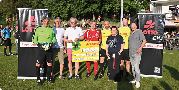 Gruppenfoto auf einem Sportplatz bei Sonnenschein:  Acht Personen stehen vorn in einer Reihe zwischen zwei schwarzen, aufrechten Werbebannern mit rotem „LOTTO“‑Logo. Die Gruppe besteht aus mehreren Erwachsenen in Sport‑ und Freizeitkleidung. In der Mitte h lt eine Person in rotem Trikot gemeinsam mit einer Person in wei em T‑Shirt einen gro en, gelben Symbol‑Scheck mit schwarzer Handschrift; auf dem Scheck ist ein Betrag von „€ 21.000“ erkennbar. Links steht eine Person in gr nem Torwarttrikot und schwarzen Torwarthandschuhen, die einen Fu ball h lt. Rechts neben der Mitte sind zwei Personen in gelben Trainingsshirts. Ganz rechts und ganz links stehen zwei Personen in l ssiger Alltagskleidung. Hinter der Gruppe ist Rasen, eine B schung mit sitzenden Zuschauern und B ume