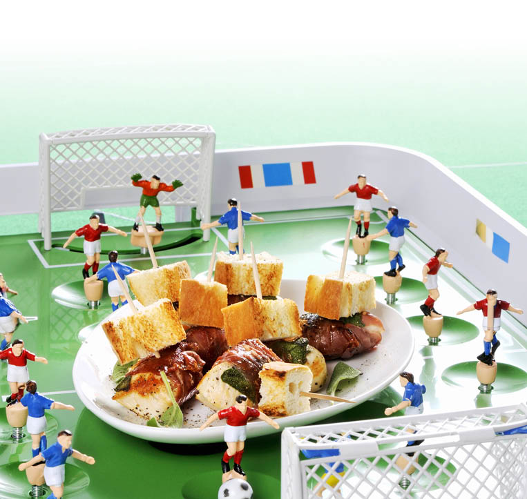 Sehr nah aufgenommene Szene eines Tischfu ballspiels:  In der Bildmitte ein wei er Teller mit einer Portion H hnchenfleischspie en, garniert mit Brotw rfeln und Salbeibl ttern auf Holzspie en. Der Teller steht auf dem gr nen Spielfeld eines Tischfu balls (Kicker). Um den Teller herum stehen kleine Holzfiguren in rot-wei en und blau-wei en Trikots, die typischen Kicker-Spieler, sowie zwei wei e Miniatur-Tore im Vorder- und Hintergrund. Helle, freundliche Farbstimmung.