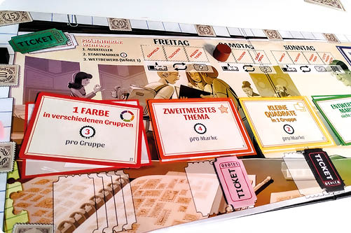 Nahaufnahme eines Brettspiel-Tableaus mit Marken und Karten: drei farbige Aufsteller zeigen Aufgaben („1 Farbe“, „Zweitmeistes Thema“, „kleine Quadrate“); oben sind Wochentage und Ticket-Felder zu sehen.