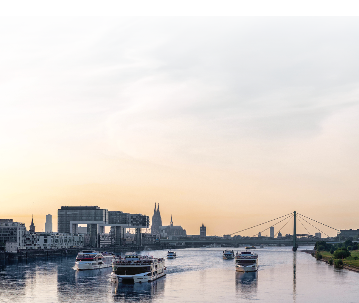 Das Foto zeigt einen weiten Blick ber den Rhein bei Sonnenuntergang in K ln. Auf dem ruhigen Wasser fahren mehrere wei e Fahrgast- und Ausflugsschiffe in beide Richtungen; ihre Spiegelungen sind auf der Wasseroberfl che erkennbar. Links am Ufer stehen moderne Geb ude, deutlich zu sehen sind die Kranh user. In der Bildmitte ragen die T rme des K lner Doms aus der Silhouette der Stadt. Rechts  berspannt eine gro e H ngebr cke den Fluss; am rechten Ufer verl uft ein schmaler Streifen mit Gr nfl chen. Der Himmel ist hell und warm get nt, von zartem Gelb bis Ros .