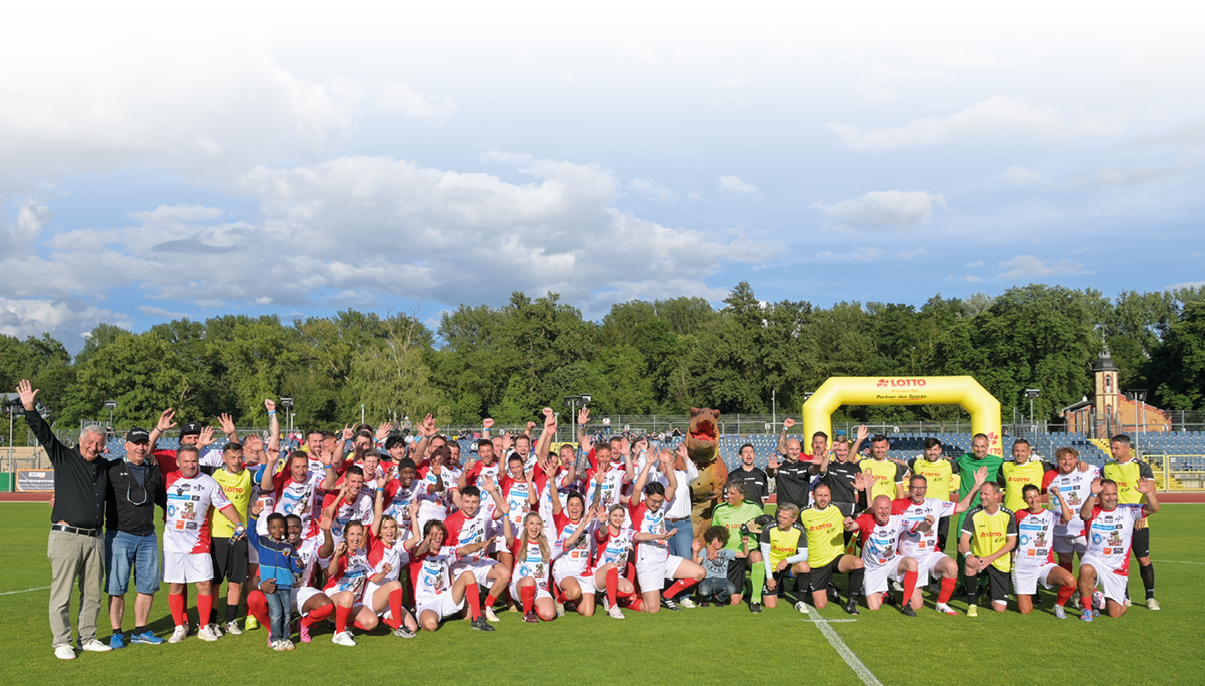 Eine gro e Gruppe von Menschen in Sportkleidung posiert fr hlich auf einem Fu ballfeld bei sonnigem Wetter vor einem gelben Torbogen mit der Aufschrift Lotto. Beschreibung von KI generiert