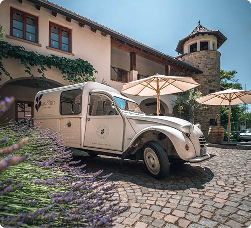 Ein wei es Oldtimer-Auto steht auf einem gepflasterten Hof vor einem Geb ude mit roten Fensterrahmen und zwei gro en Sonnenschirmen. Beschreibung von KI generiert