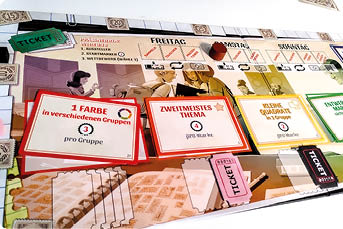 Nahaufnahme eines Brettspiel-Tableaus mit Marken und Karten: drei farbige Aufsteller zeigen Aufgaben („1 Farbe“, „Zweitmeistes Thema“, „kleine Quadrate“); oben sind Wochentage und Ticket-Felder zu sehen.