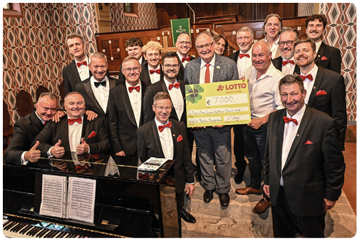 Eine Gruppe von Erwachsenen in formeller Kleidung mit Fliegen und Einsteckt chern posiert in einem Raum mit Klavier und h lt einen gro en symbolischen Scheck. Beschreibung von KI generiert