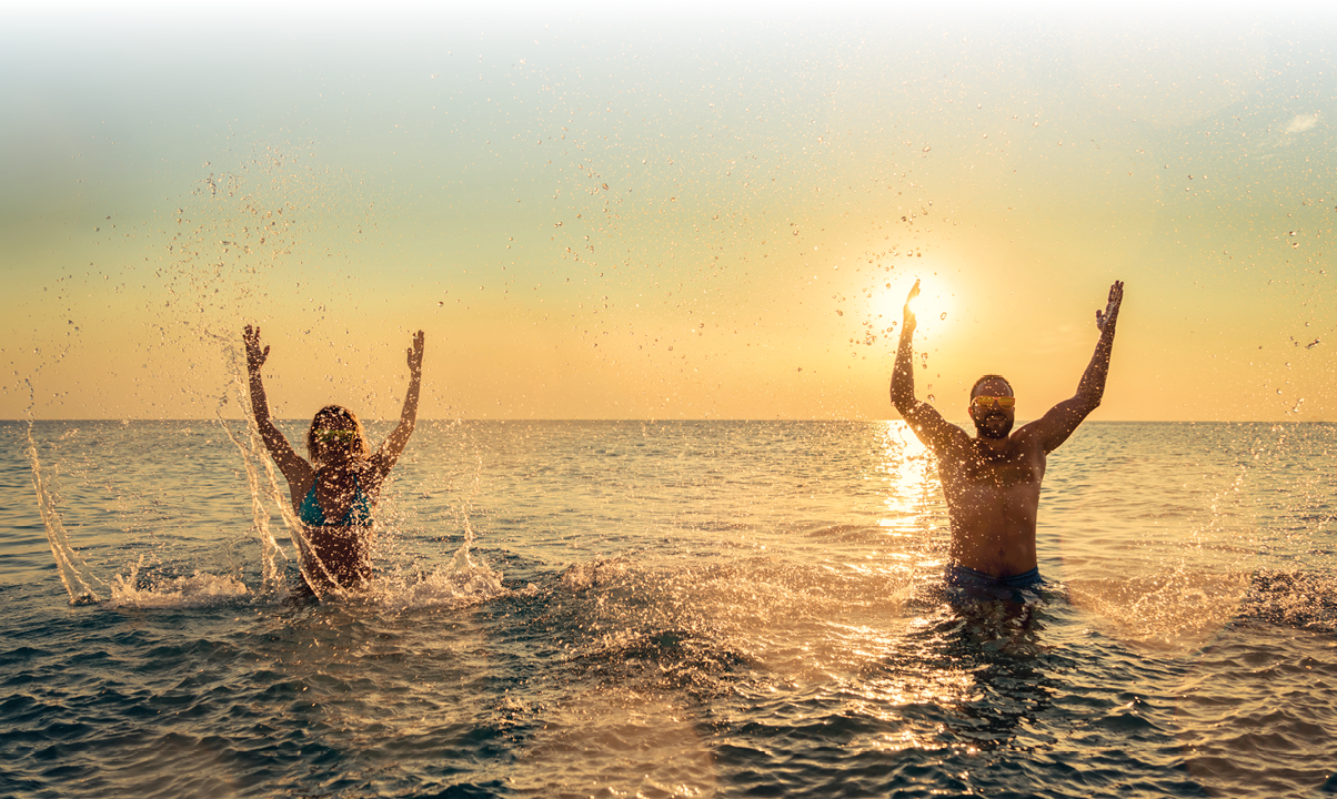 Zwei Personen springen lachend im Wasser und spritzen Wasser in die Luft bei Sonnenuntergang am Meer. Beschreibung von KI generiert