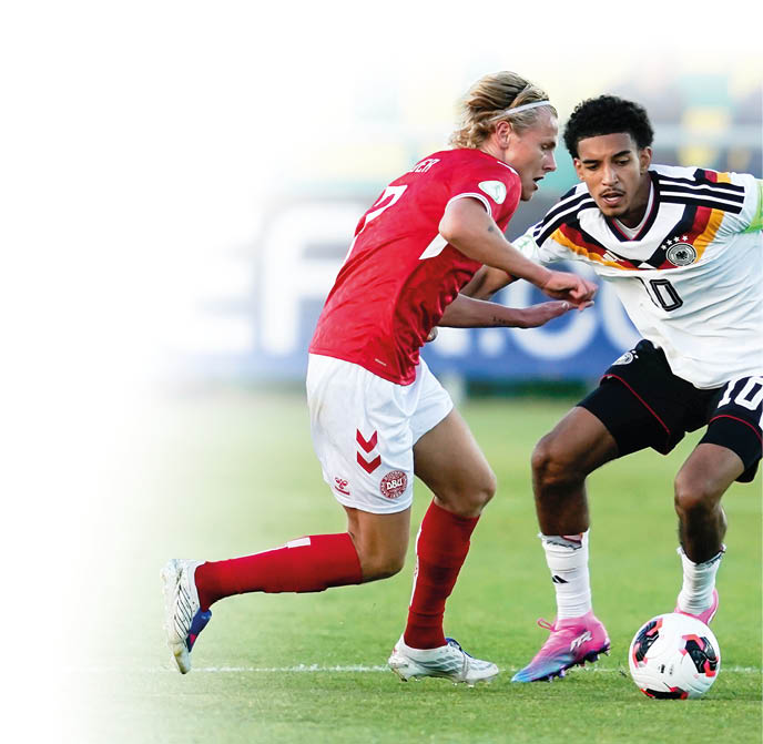 Zwei Erwachsene in Sportkleidung k mpfen auf einem Fu ballfeld um den Ball, wobei einer ein rotes Trikot und der andere ein wei es Trikot mit Kapit nsbinde tr gt. Beschreibung von KI generiert