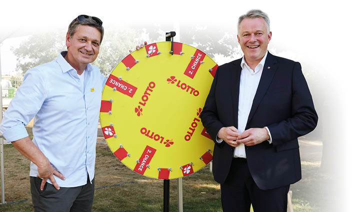 Zwei Erwachsene stehen l chelnd neben einem gro en gelben Gl cksrad mit der Aufschrift Lotto und mehreren roten Feldern mit Gewinnchancen. Beschreibung von KI generiert
