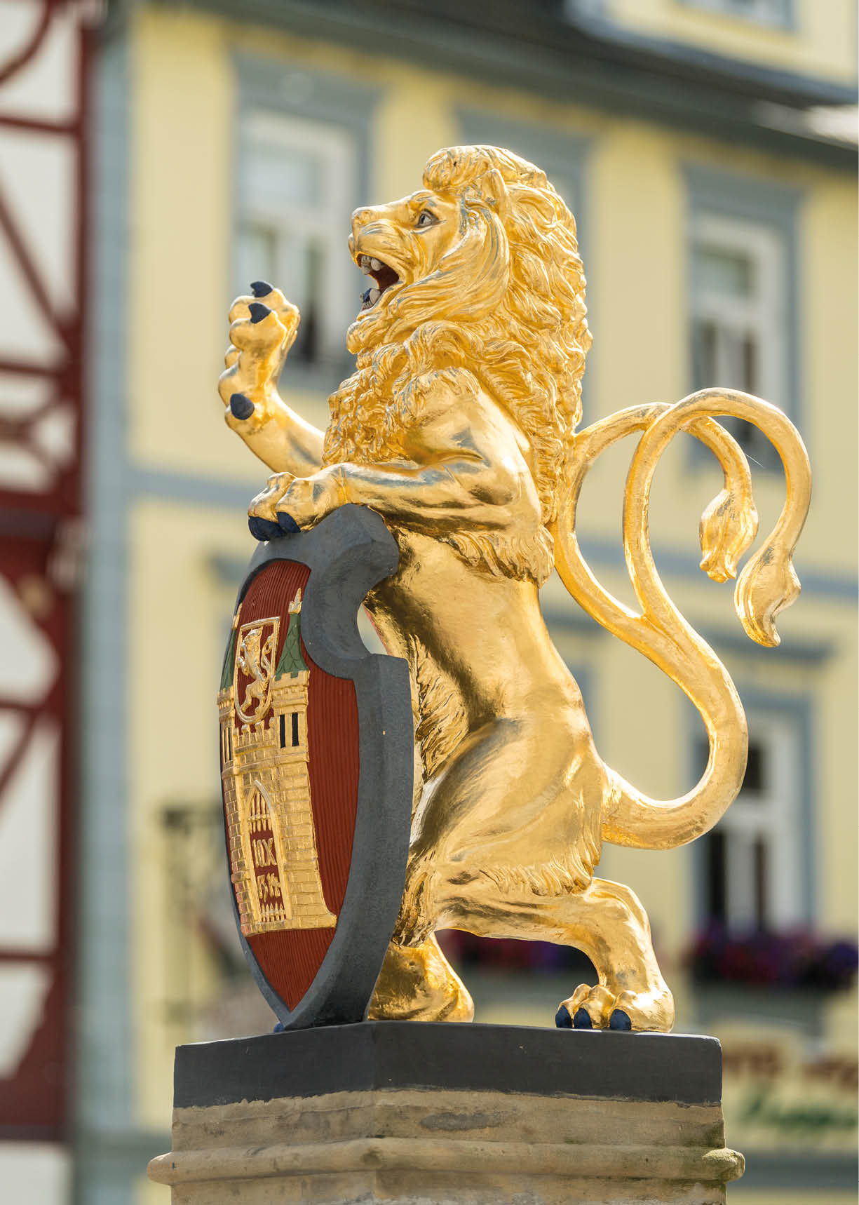 Eine goldene L wenstatue steht auf einem Sockel und h lt ein Wappenschild mit einem detaillierten Emblem vor einem unscharfen Geb ude. Beschreibung von KI generiert