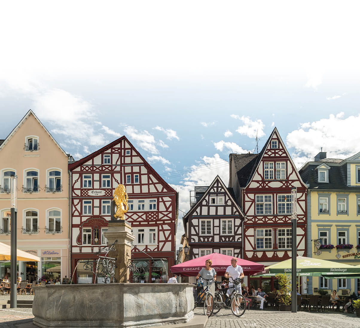 In einem belebten historischen Stadtkern stehen Fachwerkh user hinter einem Brunnen, vor dem zwei Personen mit Fahrr dern stehen und weitere Menschen in Stra encaf s sitzen. Beschreibung von KI generiert