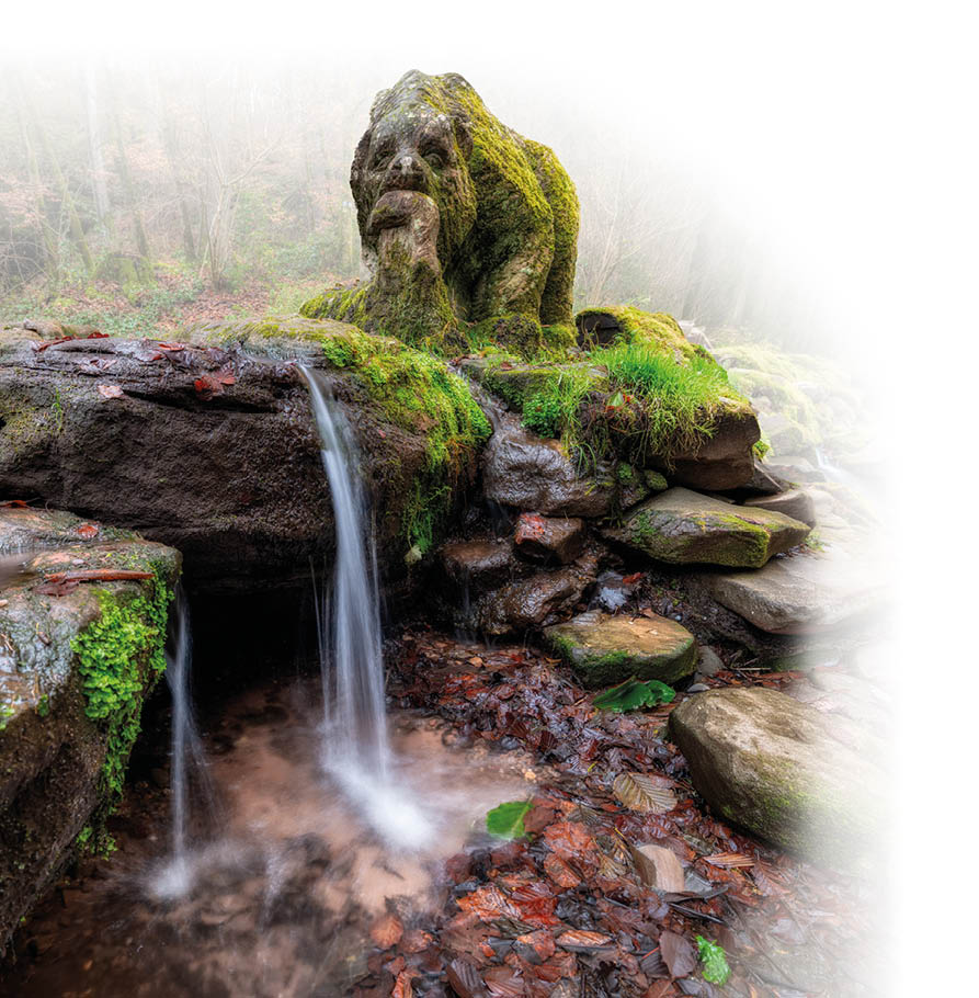 Ein kleiner Wasserfall flie t ber moosbedeckte Steine, auf denen eine moosbewachsene Skulptur in Form eines sitzenden Wesens mit einer Hand am Kinn steht.    Beschreibung von KI generiert