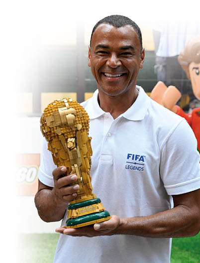 Eine l chelnde Person tr gt ein wei es Polohemd mit der Aufschrift FIFA LEGENDS und h lt eine aus LEGO-Steinen gebaute Nachbildung des FIFA-Weltpokals. Beschreibung von KI generiert