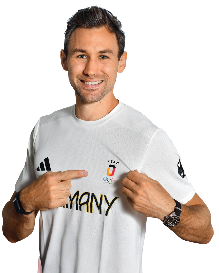 Eine Person tr gt ein wei es Sporttrikot mit der Aufschrift Germany und zeigt mit beiden H nden auf das Logo des Teams Deutschland f r die Olympischen Spiele. Beschreibung von KI generiert