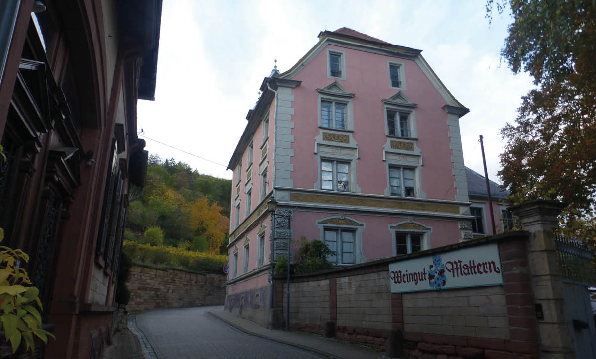 Ein historisches, rosa gestrichenes Geb ude mit grauen Fensterrahmen steht an einer kurvigen Stra e, daneben ein Schild mit der Aufschrift „Weingut Mattern“. Beschreibung von KI generiert