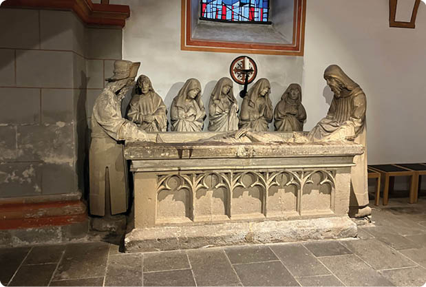 Eine steinerne Skulpturengruppe zeigt mehrere Personen in historischen Gew ndern, die um einen Sarkophag in einem Raum mit einem bunten Glasfenster versammelt sind. Beschreibung von KI generiert