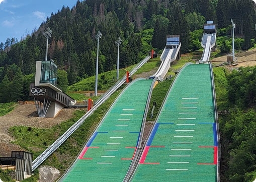Eine gr ne Skisprungschanze mit Start- und Landebereich umgeben von B umen und einer Wiese unter einem bew lkten Himmel. Beschreibung von KI generiert