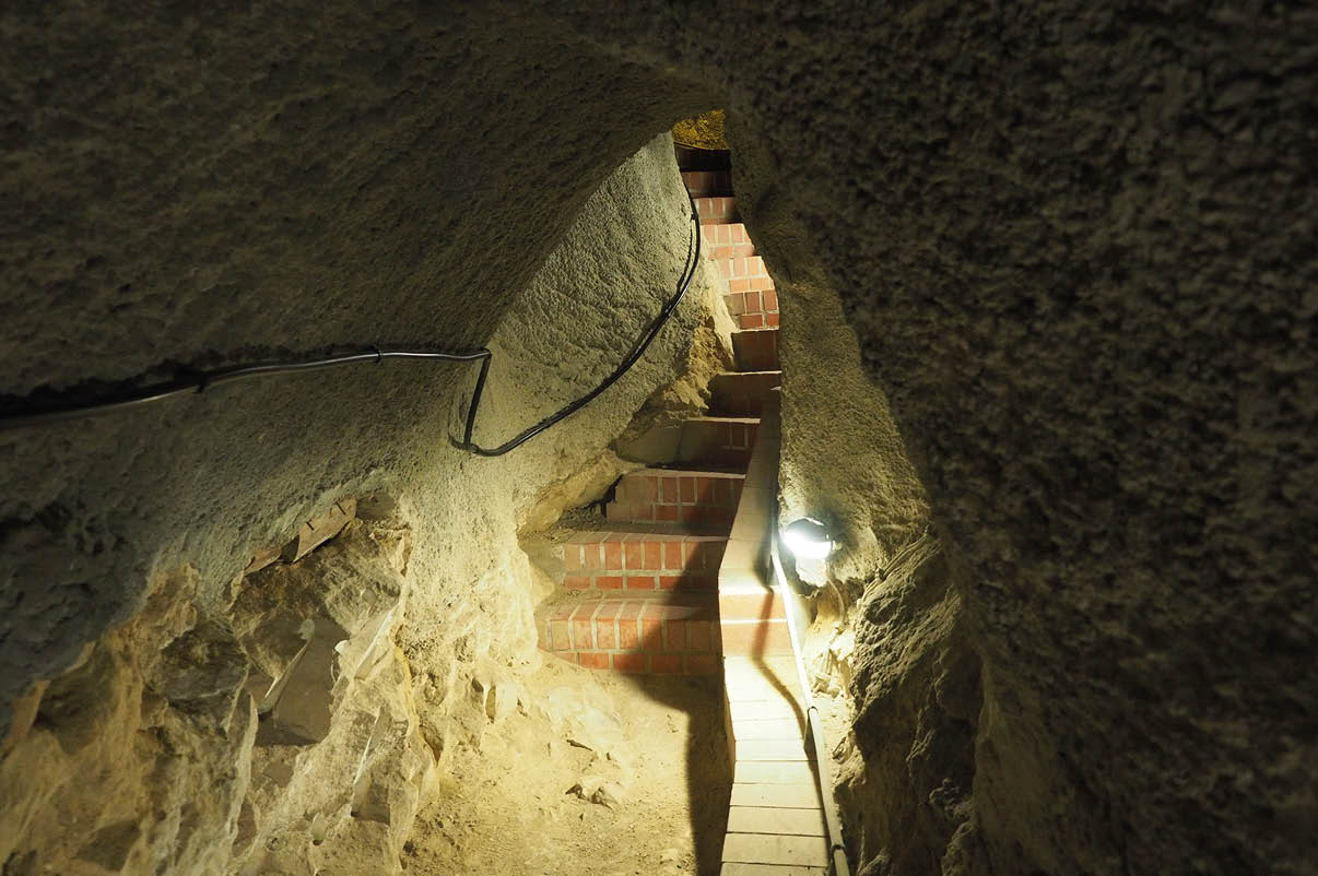 Ein schmaler, beleuchteter Tunnel mit unregelm igen W nden f hrt zu einer kleinen Treppe aus roten Ziegeln.  Beschreibung von KI generiert