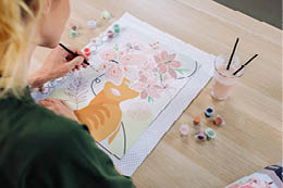 Person malt mit Pinsel ein Bild einer Katze und Blumen auf Papier mit mehreren kleinen Farbt pfchen und einem Getr nk auf dem Tisch