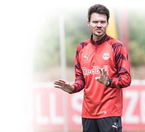 Eine Person mit kurzem Haar tr gt ein rotes Sporttrikot mit Red Bull-Logo und gestikuliert auf einem Fu ballfeld vor einem unscharfen Hintergrund mit der Aufschrift Salzburg. Beschreibung von KI generiert