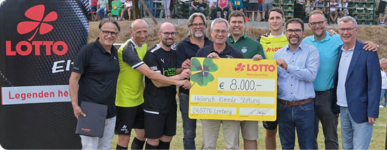 Eine Gruppe von Erwachsenen steht auf einem Rasen vor zwei Bannern mit dem Lotto-Logo und h lt gemeinsam einen gro en symbolischen Scheck, w hrend im Hintergrund Zuschauer auf Trib nen sitzen. Beschreibung von KI generiert