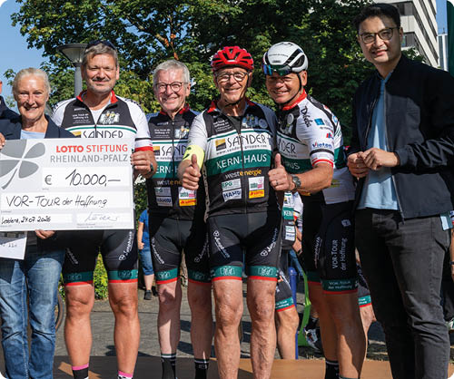 Eine Gruppe von Erwachsenen in Sportbekleidung und Freizeitkleidung steht auf einer B hne im Freien und h lt einen gro en symbolischen Scheck bei einer Veranstaltung mit Bannern im Hintergrund. Beschreibung von KI generiert