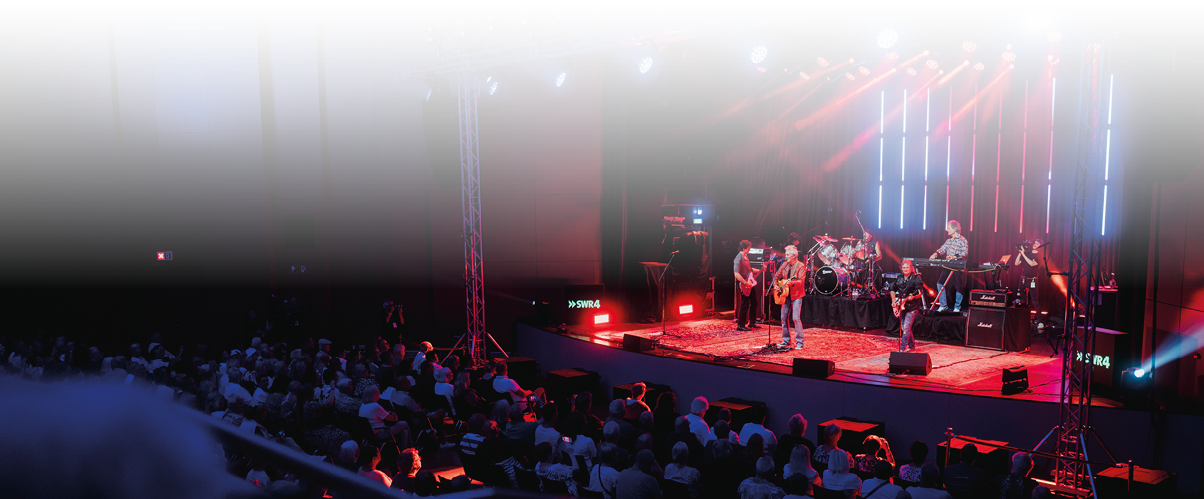 Eine Band spielt auf einer B hne mit bunten Lichtern vor einem Publikum in einem dunklen Konzertsaal. Beschreibung von KI generiert