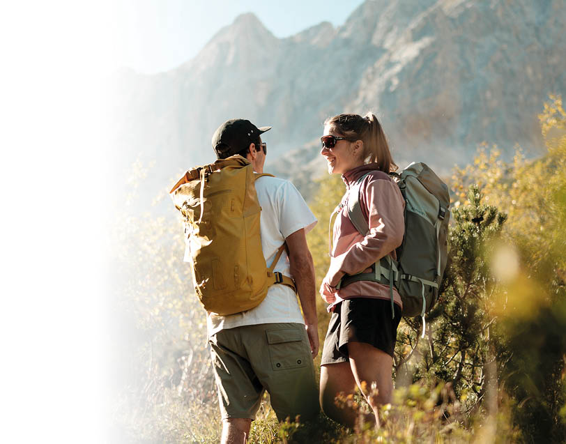 Zwei Personen mit Rucks cken und Outdoor-Kleidung unterhalten sich beim Wandern in einer bergigen Landschaft bei sonnigem Wetter. Beschreibung von KI generiert