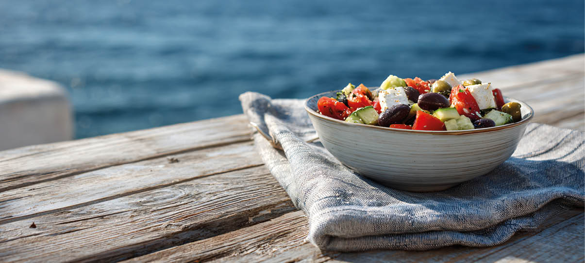 Eine Sch ssel mit einem bunten Salat aus Tomaten, Gurken, Oliven und Feta steht auf einem grauen Tuch auf einem h lzernen Tisch mit Blick auf das Meer im Hintergrund. Beschreibung von KI generiert