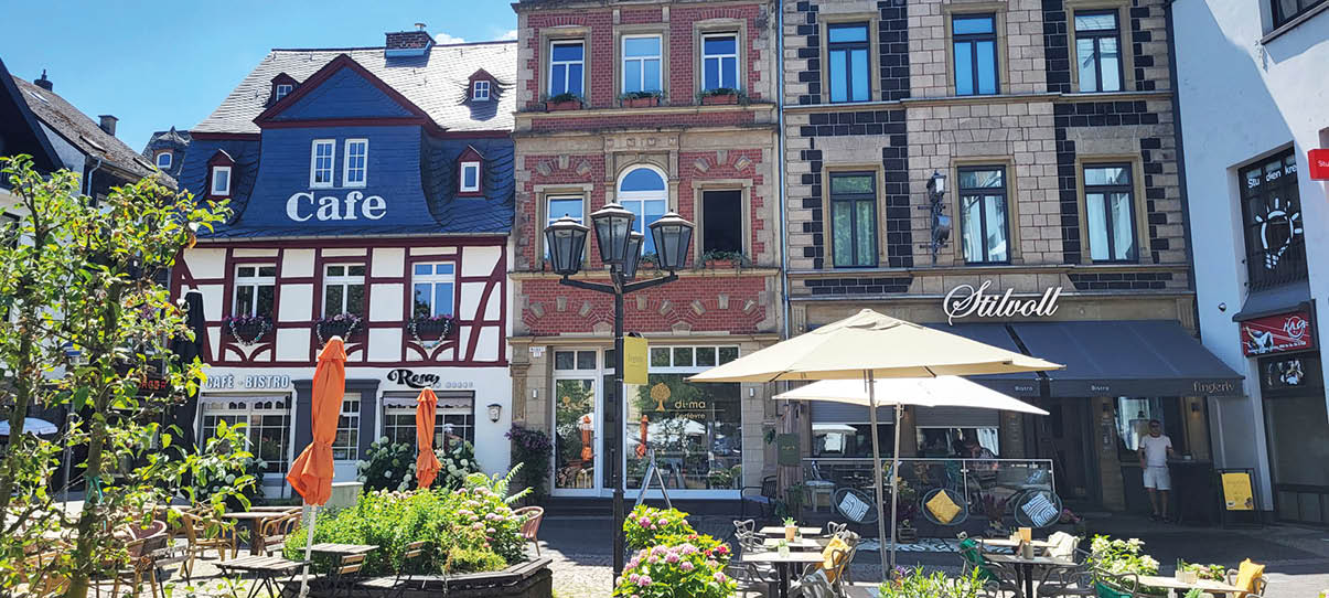 Ein sonniger Stra encaf bereich mit Tischen, St hlen, Sonnenschirmen und Pflanzen vor historischen Fachwerk- und Steingeb uden mit Caf s und Gesch ften. Beschreibung von KI generiert