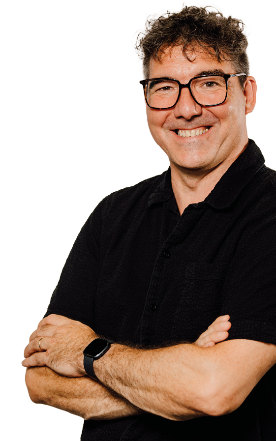 Eine l chelnde Person mit lockigem Haar, Brille und schwarzem Hemd steht mit verschr nkten Armen vor einem hellen Hintergrund. Beschreibung von KI generiert
