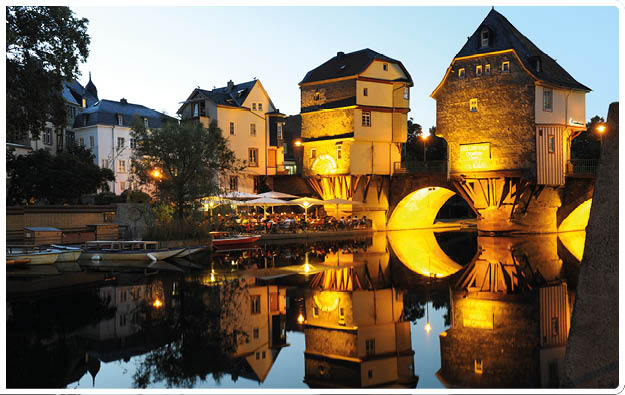 Abendliche Szene mit historischen Fachwerkh usern auf einem beleuchteten Steg ber einem ruhigen Fluss, der die Geb ude und Lichter klar reflektiert.  Beschreibung von KI generiert