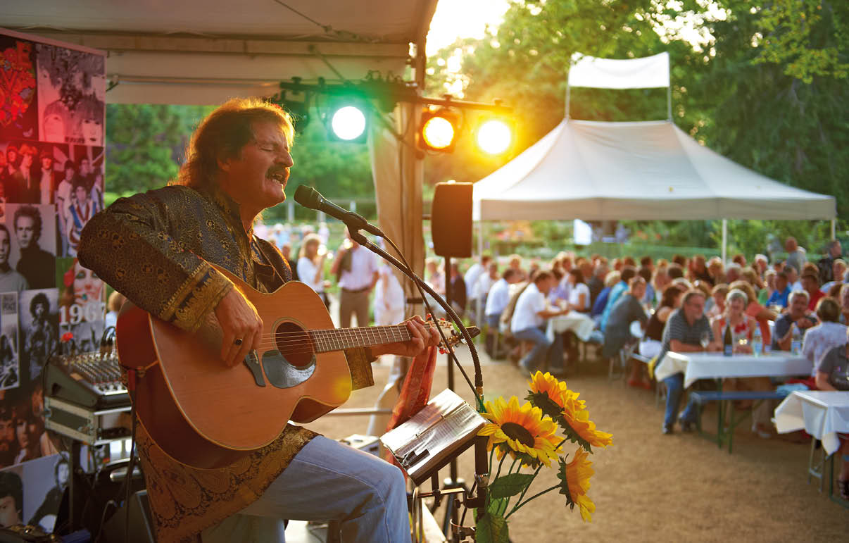Eine Person spielt Gitarre und singt in einem Zelt vor einer gro en Gruppe von sitzenden Menschen bei Sonnenuntergang. Beschreibung von KI generiert
