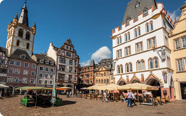 Historischer Marktplatz mit Fachwerkh usern, einem Turm und Menschen, die bei Sonnenschein an Tischen unter Sonnenschirmen sitzen und spazieren. Beschreibung von KI generiert
