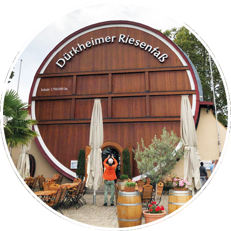 Vor einem gro en h lzernen Fass mit der Aufschrift D rkheimer Riesenfass stehen mehrere Menschen und Tische mit St hlen sowie zwei geschlossene Sonnenschirme. Beschreibung von KI generiert